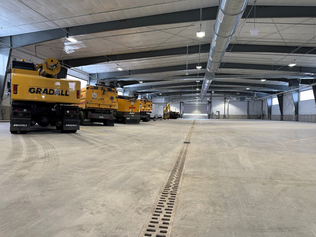 Wide indoor industrial warehouse with yellow Gradall construction machines parked along the left wall and large overhead ducts. A central floor trench runs down the middle.