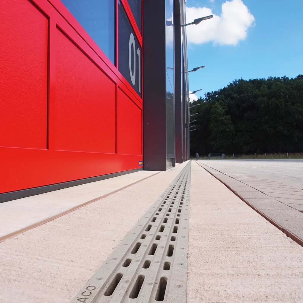 Low-angle view of a bright red building facade with glass panels, beside a long metal drainage grate along a paved walkway under a blue sky.