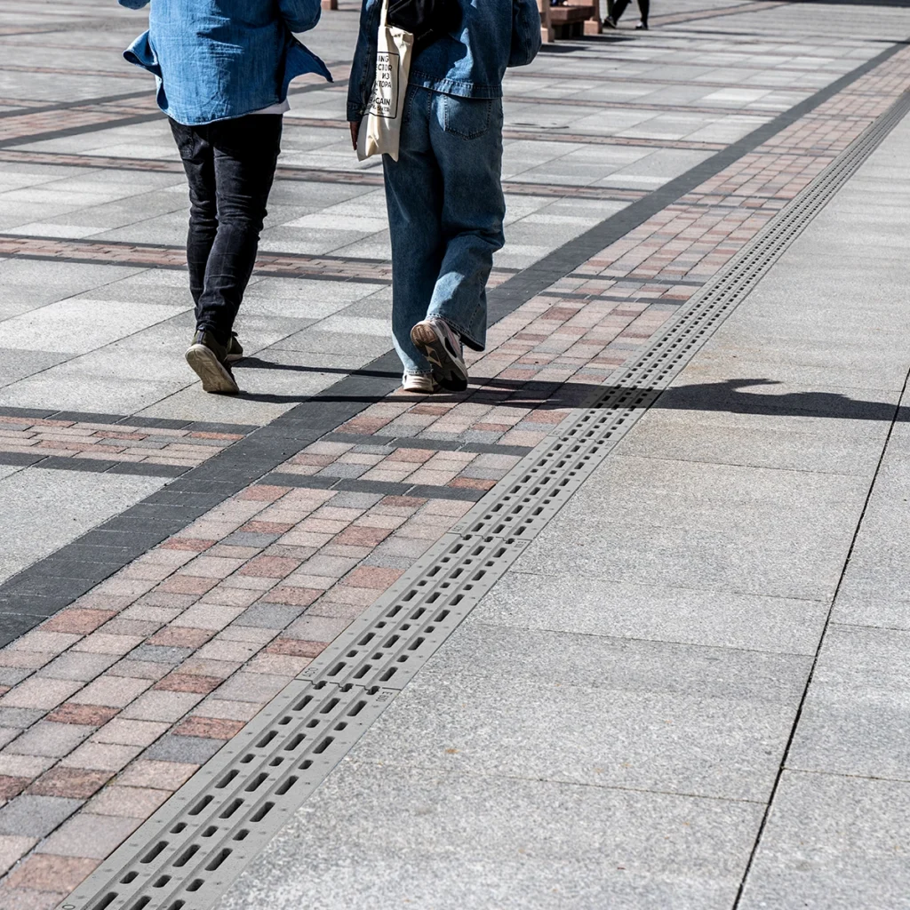 Two people walk away on a patterned plaza; one carries a tote bag, the other wears jeans and sneakers, with a long metal drainage grate nearby.