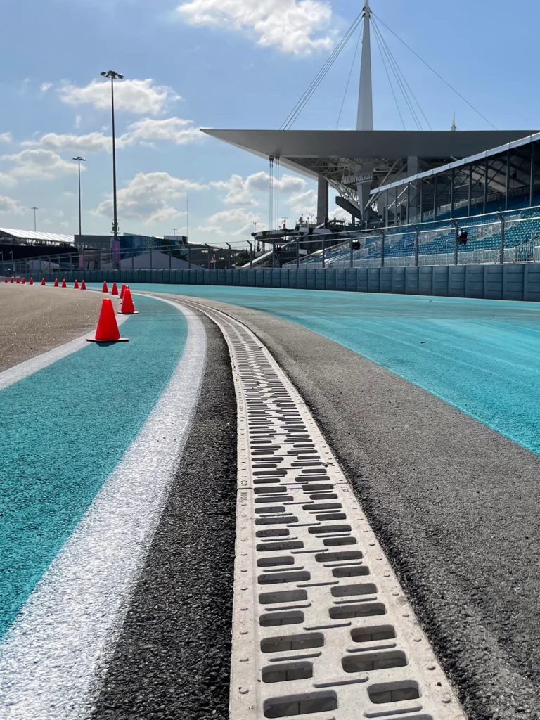 Race track curve with teal run-off area, orange safety cones along the edge, and a modern grandstand under a blue sky.