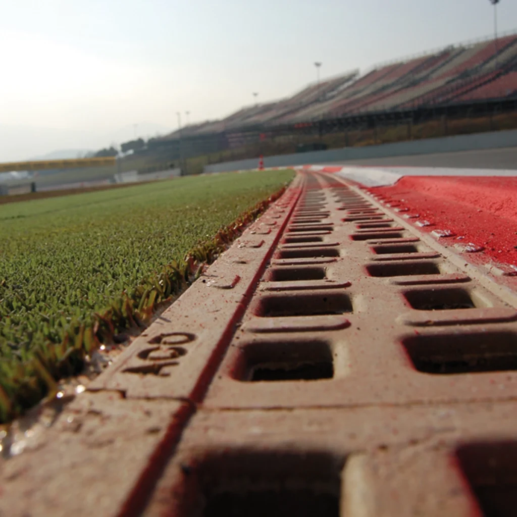Low-angle close-up of a red, perforated curb along a race track with green grass to the left and empty stadium seating in the distance on a sunny day.