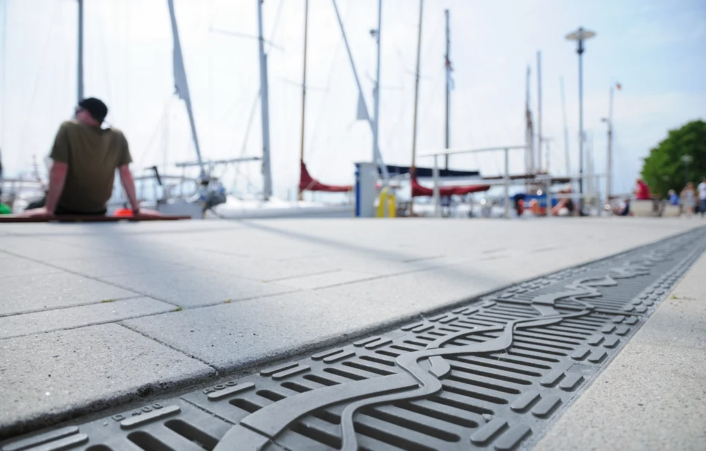 Low-angle view of a drain grate along a boardwalk with moored sailboats and a man sitting in the background at left.