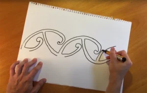 Person drawing a decorative looping pattern on a white sketchbook on a wooden table.