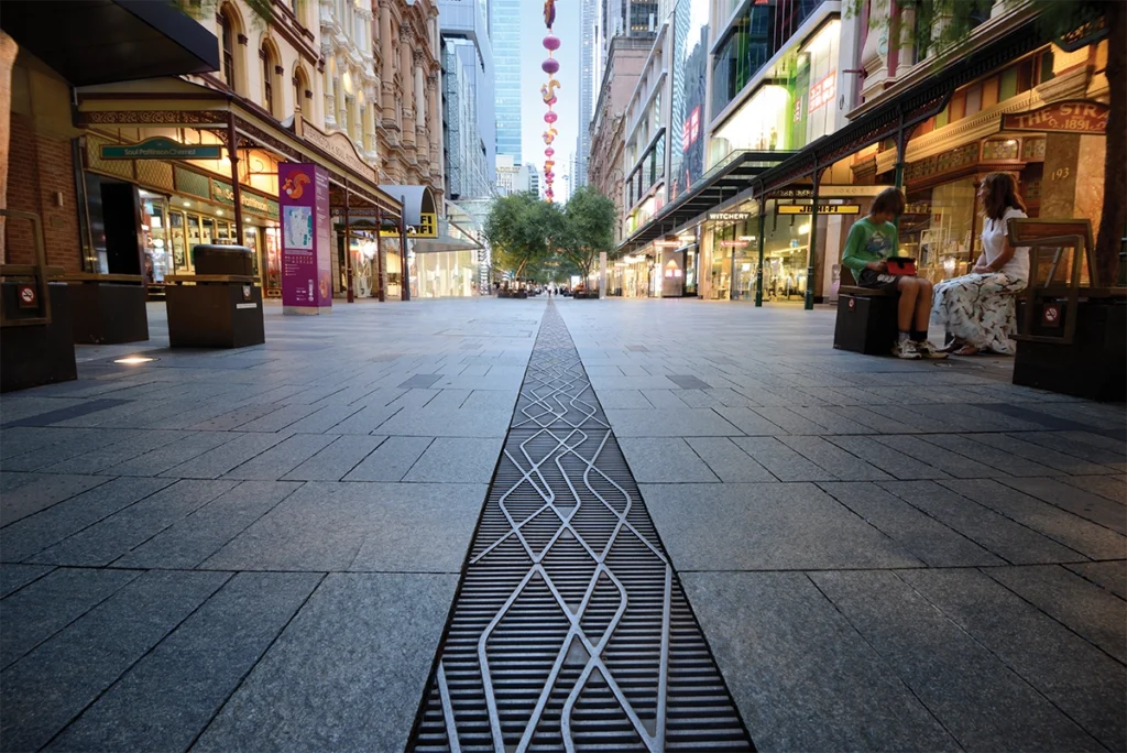 Ground-level view of a wide city pedestrian mall with a patterned metal grate running down the center, shops and storefronts on both sides, and two people sitting on benches at the right.