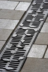 Metal street drain grate with scattered small leaves on top, set in a gray tiled sidewalk edge.
