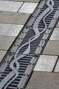 Close-up of a metal trench drain cover with a decorative wavy pattern on a paved sidewalk.