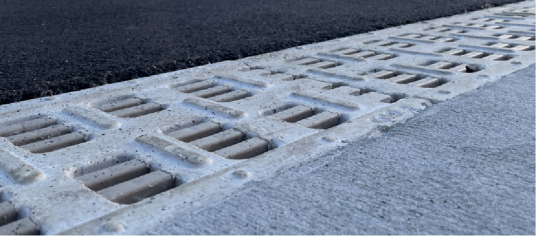 Close-up of a concrete sidewalk edge with a metal drainage grate running along the curb edge.