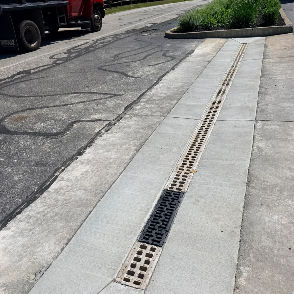 Concrete sidewalk with a long drainage grate running parallel to the curb, and a patched, rough asphalt road nearby; a truck is parked on the street in the background.