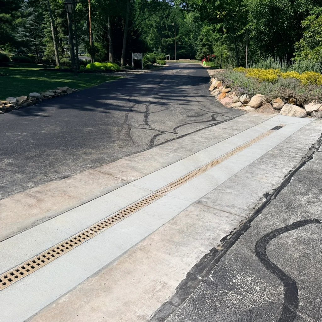 Asphalt driveway with concrete curb and a yellow drainage grate, bordered by rocks and green shrubbery on a sunny day.