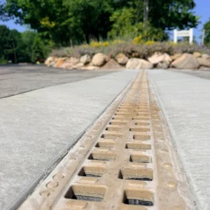 Close-up, ground-level view of a beige tactile paving strip running along a concrete sidewalk, with a metal trench drain in the center and rocks and greenery in the background.