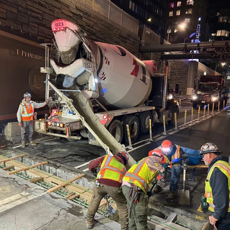 Nighttime construction scene with workers in high-visibility vests; a cement mixer truck and freshly poured trench repair on a city street, with steel rebar and wooden forms nearby