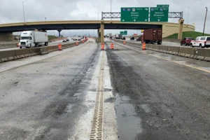 Highway with lane closures under an overpass, wet pavement, and orange traffic cones; green overhead signs point to routes toward South Bend, Chicago, and Milwaukee.
